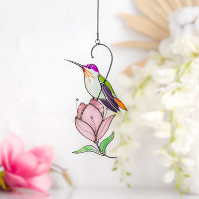 Stained glass hummingbird with purple head and green wings, perched on a soft pink magnolia flower.