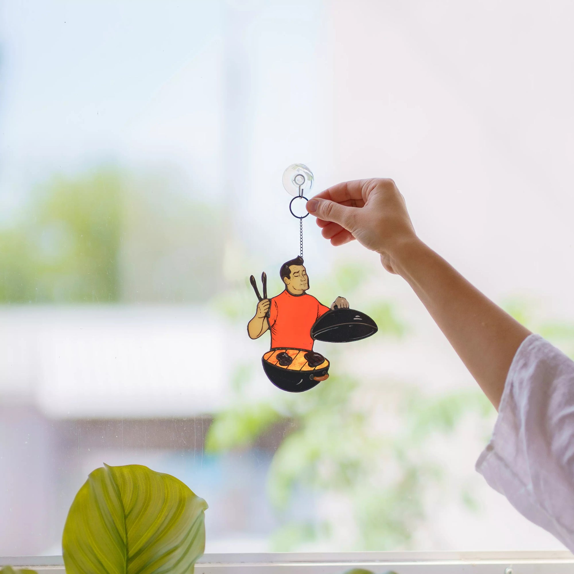 Hand holding a window hanging of a person cooking over a grill, with a blurred outdoor background.