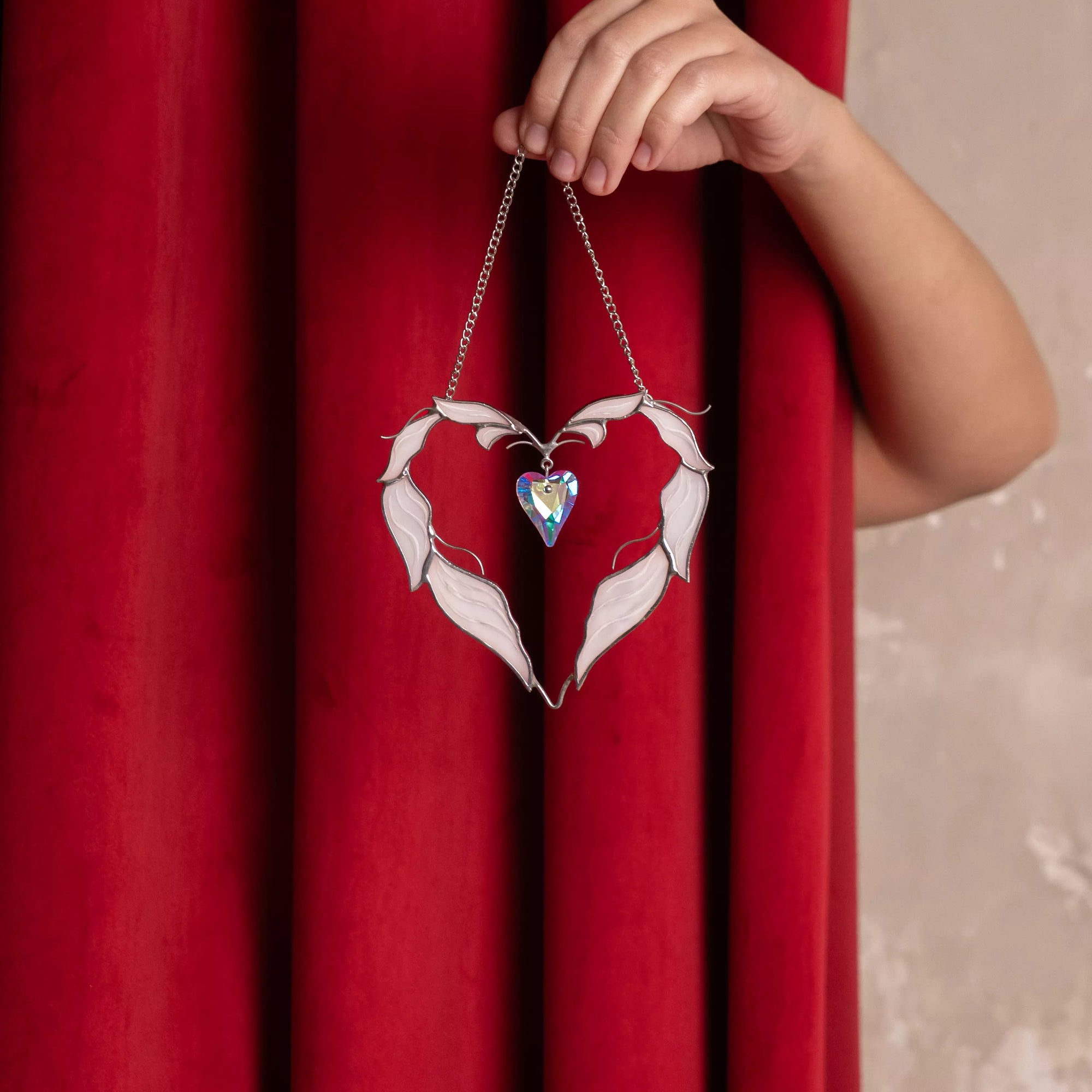 Winged heart stained glass suncatcher displayed against a rich red curtain background.