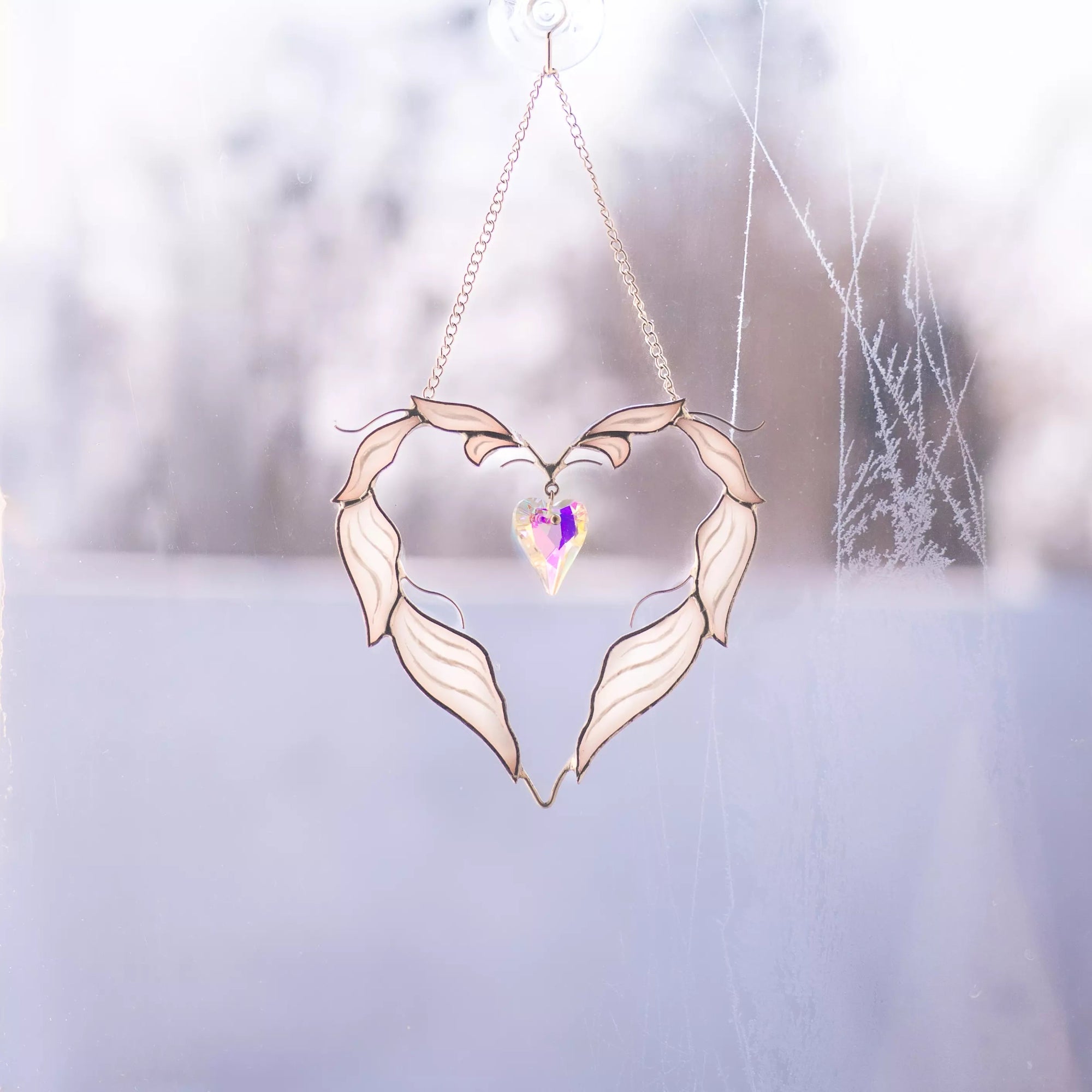 Winged heart stained glass suncatcher attached to a window with a suction cup, glowing in natural daylight.