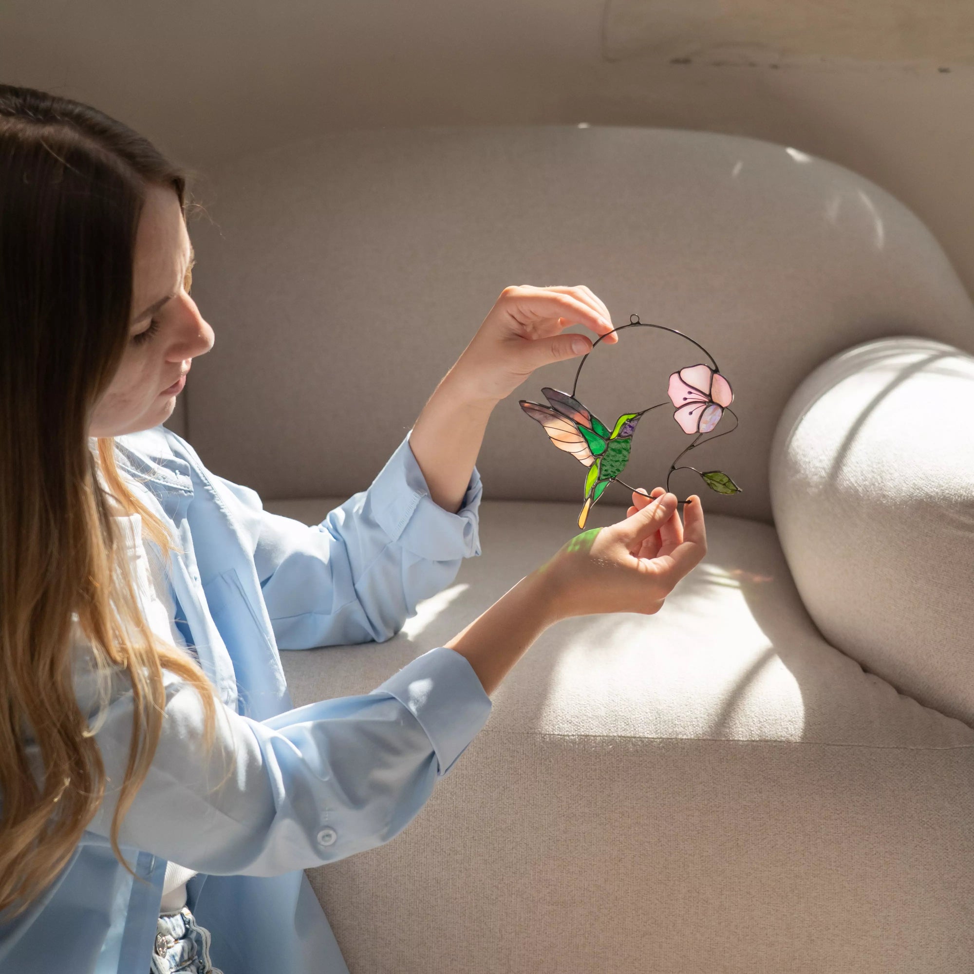 Woman holding a decorative window hanging with a hummingbird and lotus  design in a softly lit room.