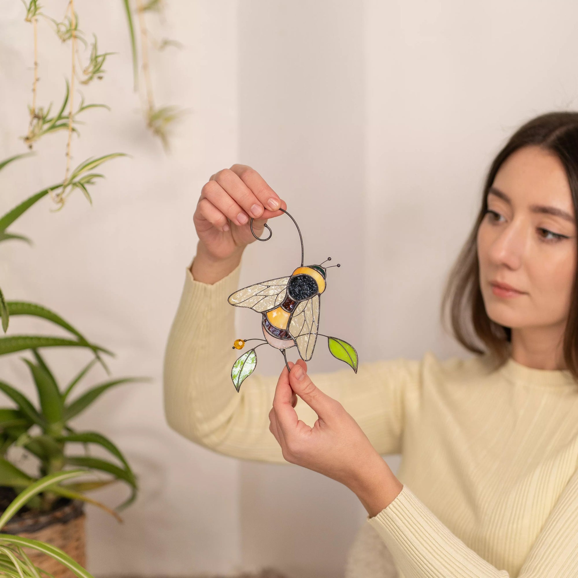 Woman holding a stained glass bumble bee suncatcher in front of a white wall with plants.