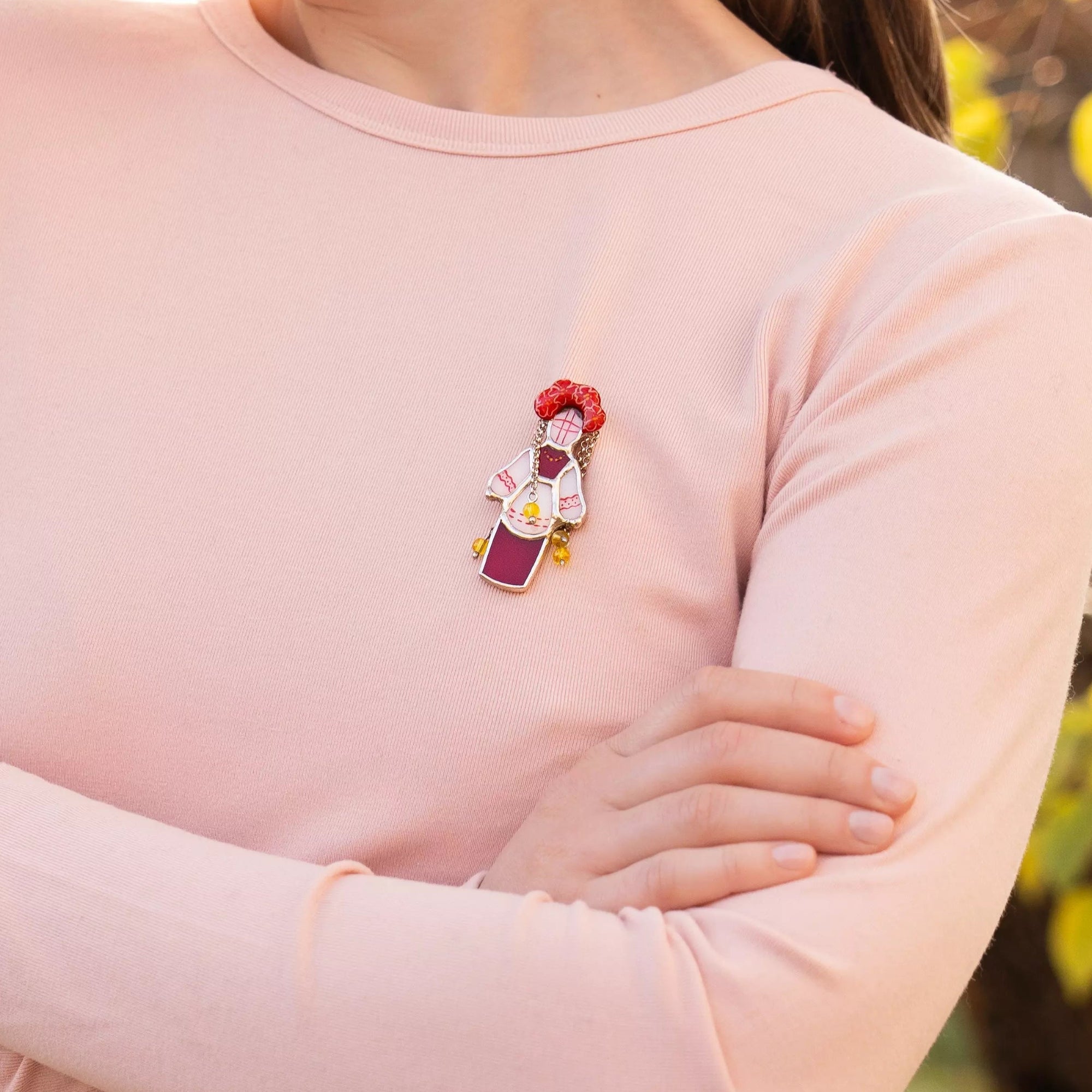 Woman wearing a pink sweater with a small red and yellow ukrainian traditional guardian doll brooch, standing outdoors with autumn leaves in the background.