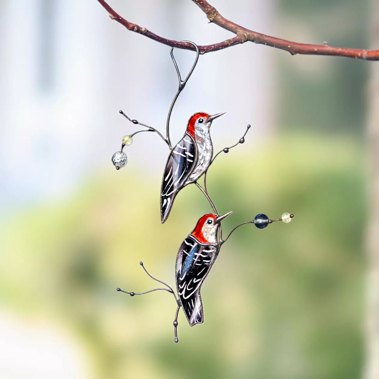 A lifestyle shot of the woodpecker suncatcher hanging from a natural tree branch outdoors, with a soft blurred garden background.