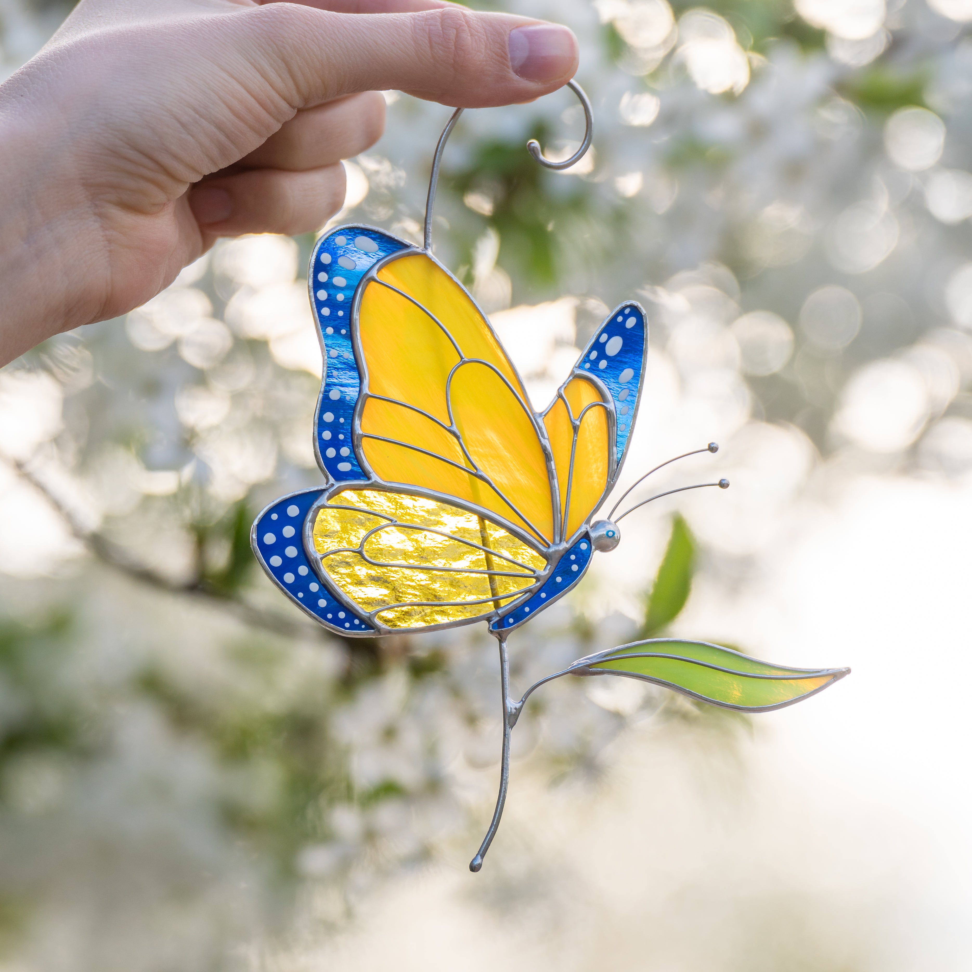 Hand holding stained glass butterfly suncatcher outdoors, bright yellow wings with blue edges.