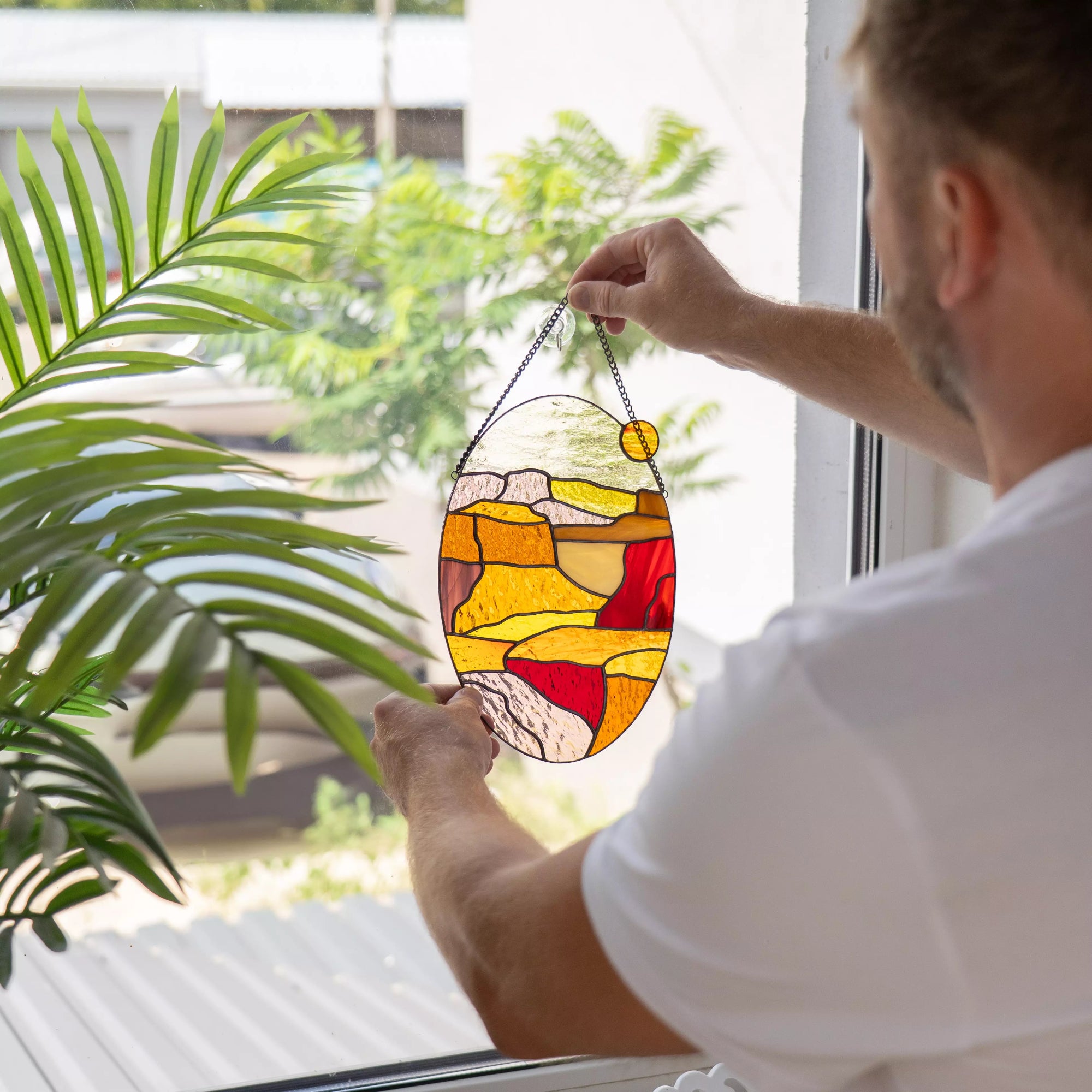 Man holding oval stained glass landscape panel up to bright window with greenery behind.