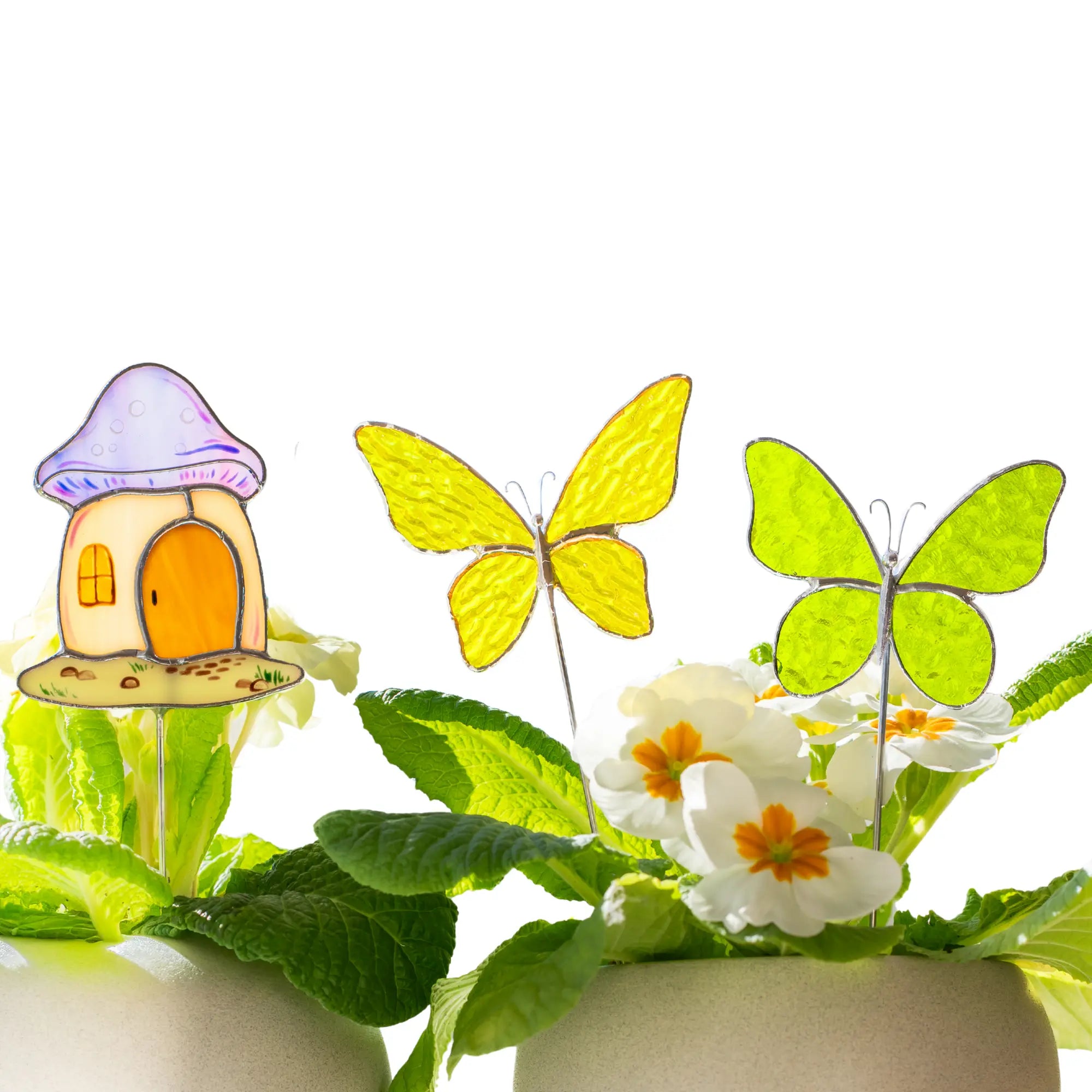 Three stained glass plant stakes - a mushroom house, a yellow butterfly, and a green butterfly - standing in white pots with green leaves against a clean white background.
