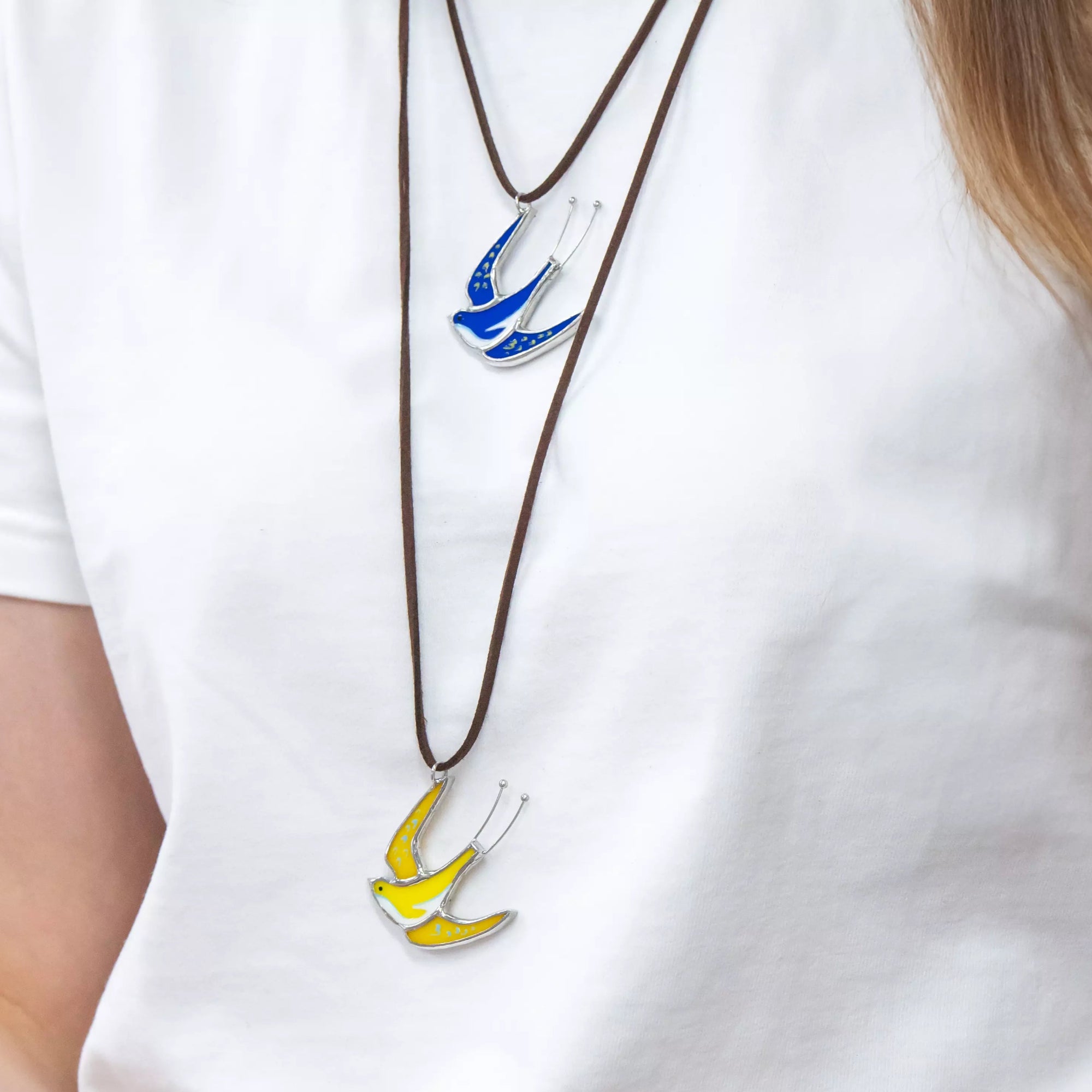 Two swallow necklaces in yellow and blue worn with a white t-shirt, hand holding a piece of stained glass.