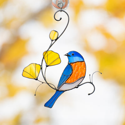 Stained glass bluebird with orange tummy and yellow flowers, hanging on a window with autumn background