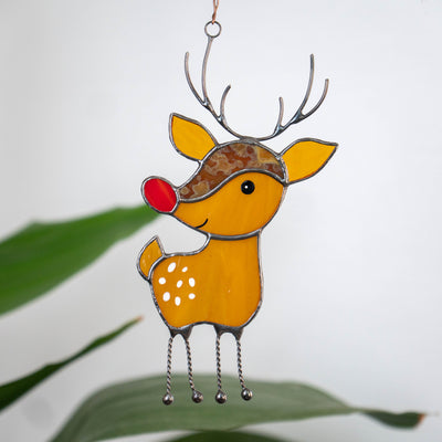 A stained glass reindeer ornament with a red nose and dotted yellow body hangs in front of a white background with green leaves.