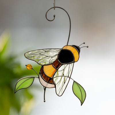 Glass bee ornament with bright amber wings and green leaves, softly lit from behind.