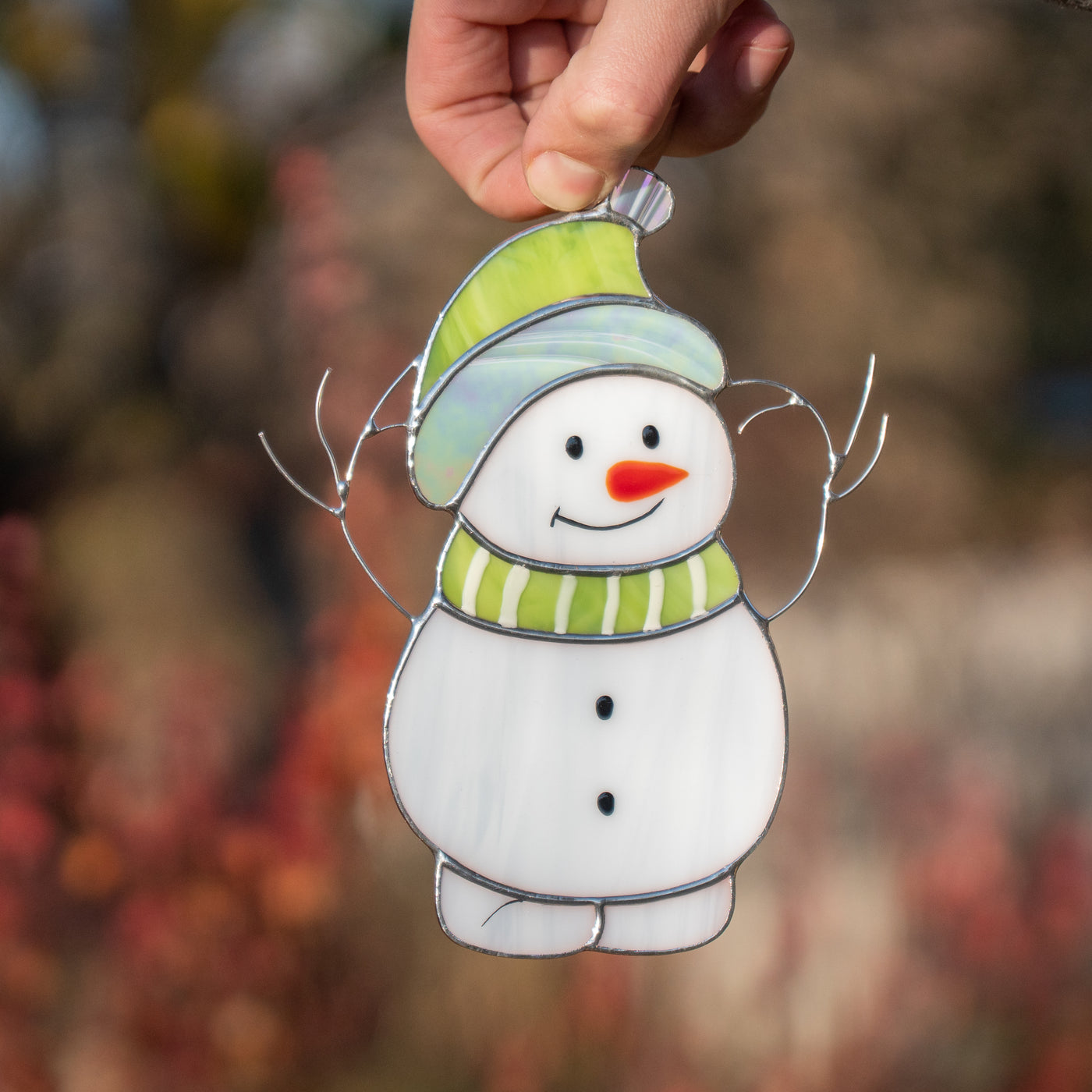 A person holding a smiling stained glass snowman with green hat and striped scarf against blurred natural background