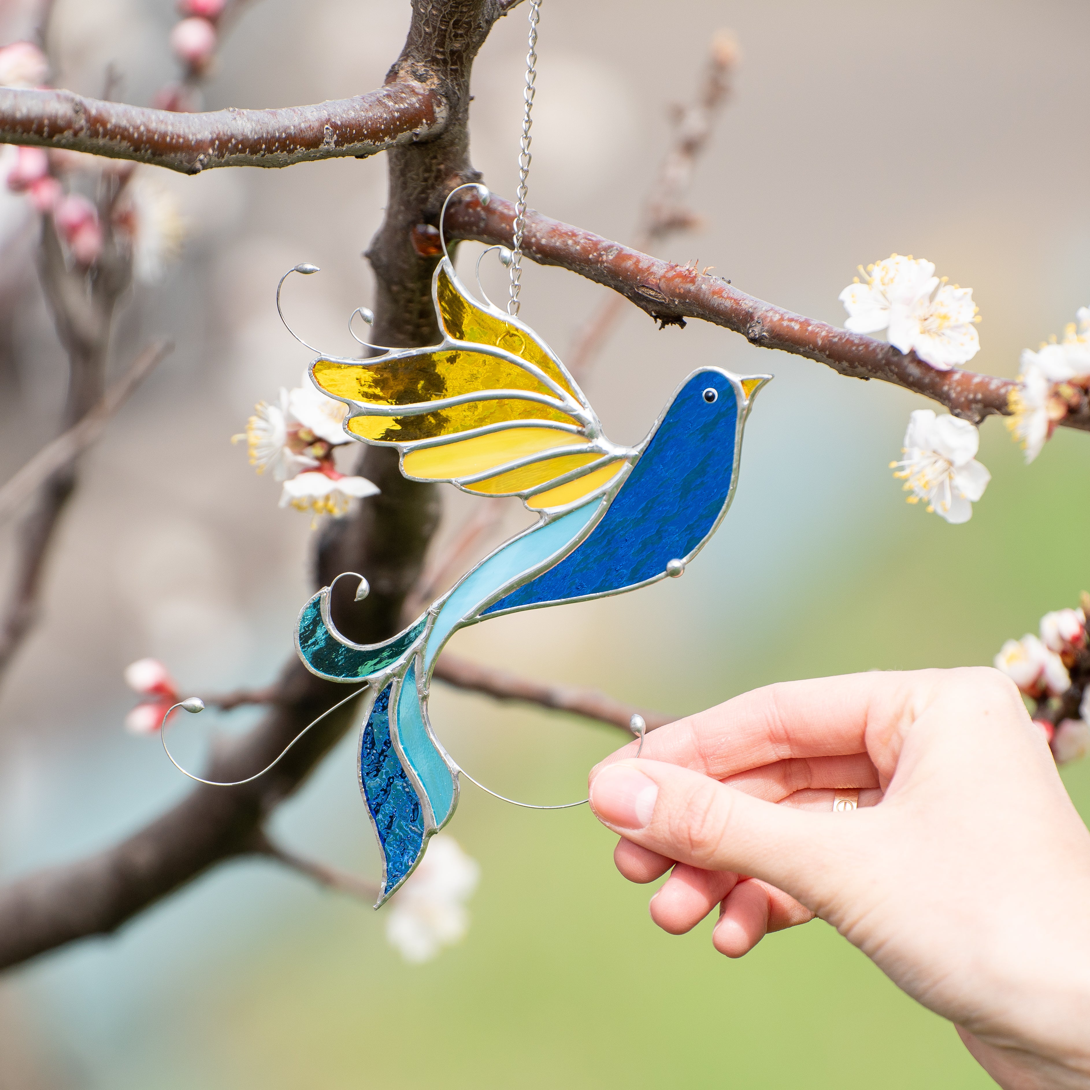 Hand holding stained glass bird with spiraled tail near blooming branch, soft background.