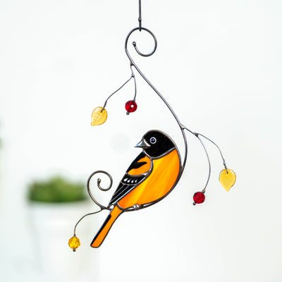 Looking left Baltimore oriole sitting on the branch with leaves and berries window hanging of stained glass