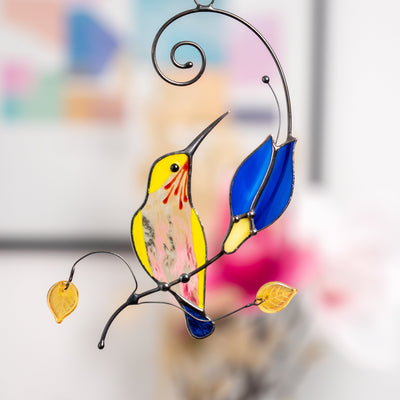 Stained glass yellow hummingbird with a blue tail sitting on the branch with the blue flower suncatcher