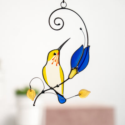 Yellow hummingbird with the blue tail on the branch with blue flower window hanging of stained glass