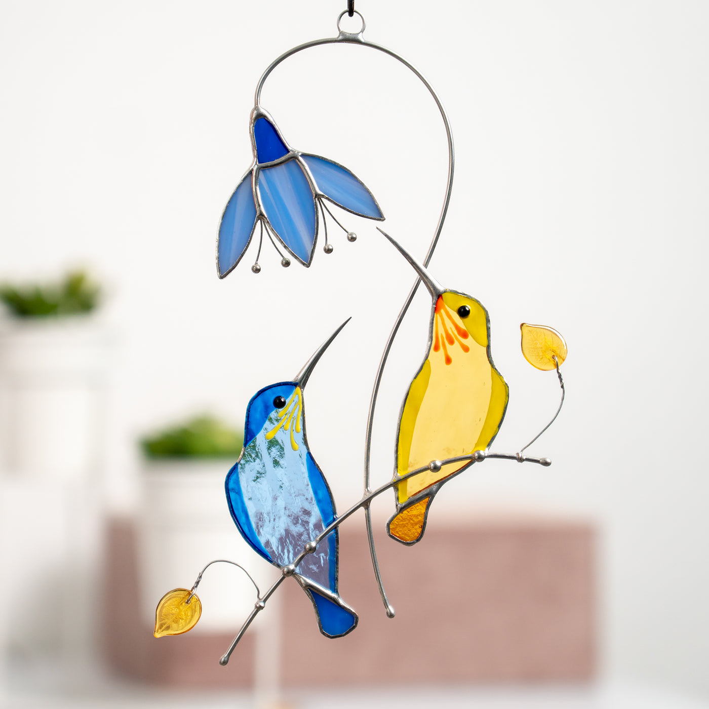 Couple of stained glass blue and yellow hummingbirds sitting on the branch with the blue flower above suncatcher