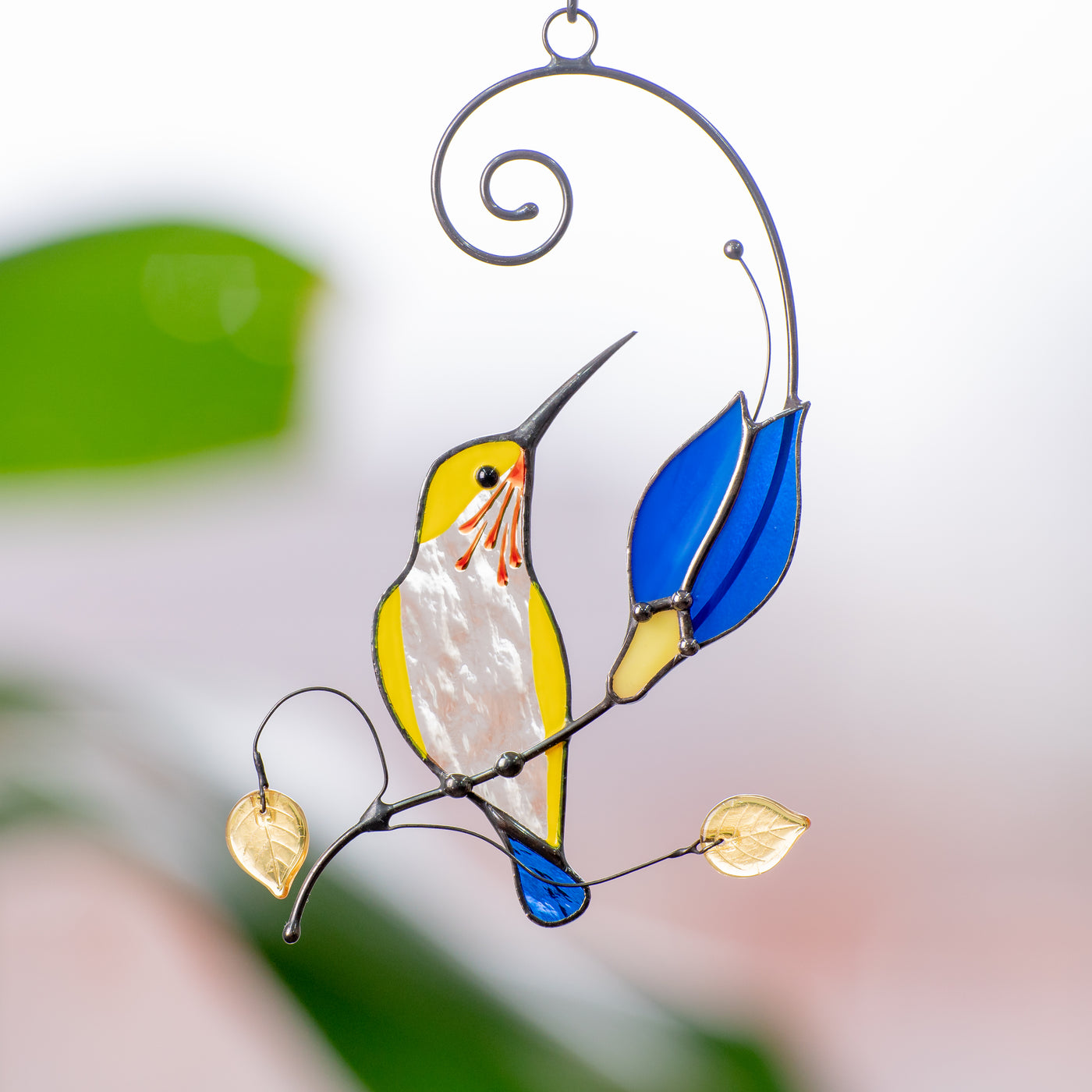 Stained glass window hanging of a yellow hummingbird with a blue tail sitting on the branch with the blue flower