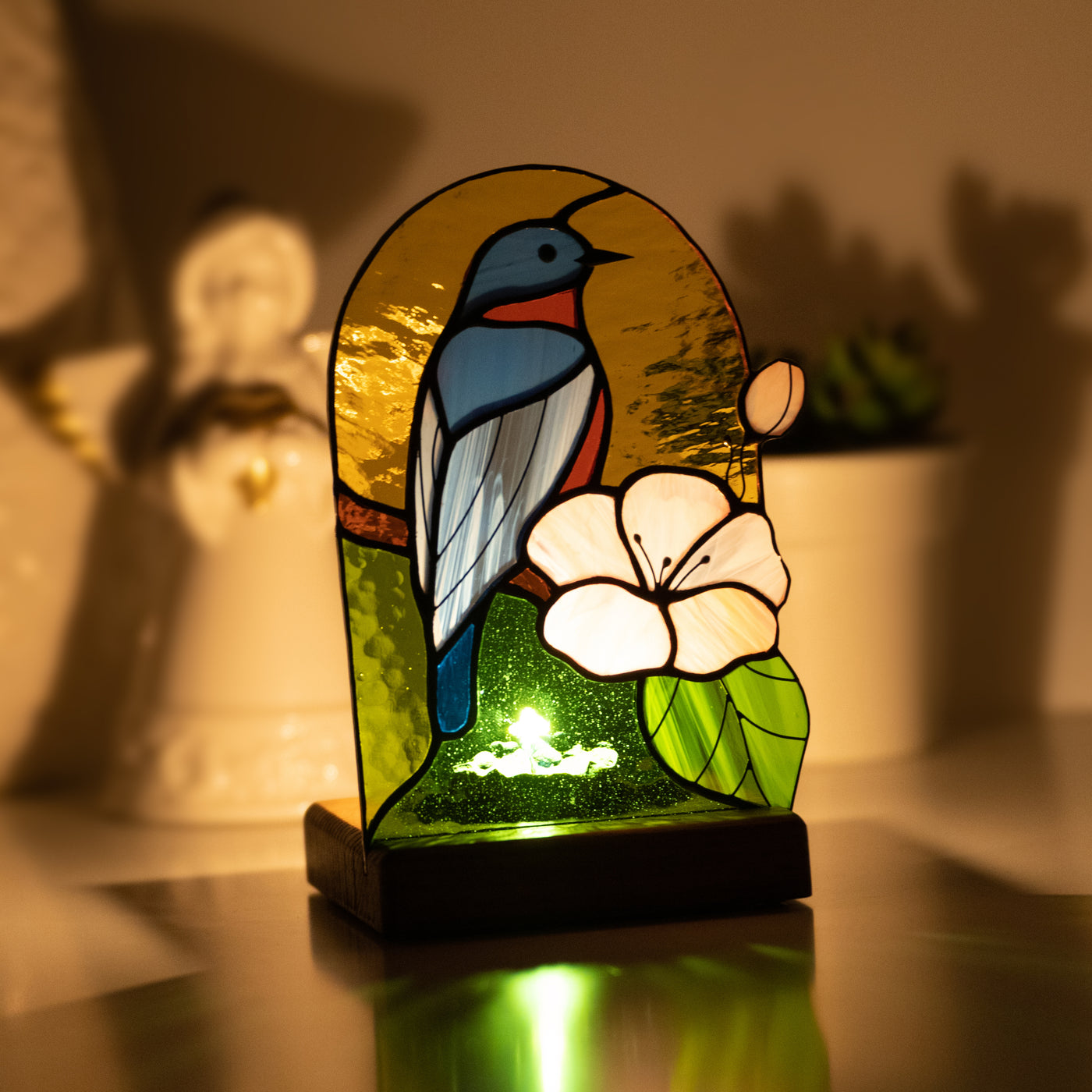 Stained glass panel candle-lit panel in a wooden base depicting bluebird and pink flower in the dark