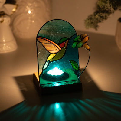 Stained glass candle-lit panel in a wooden base depicting a flying hummingbird and yellow flower in the dark
