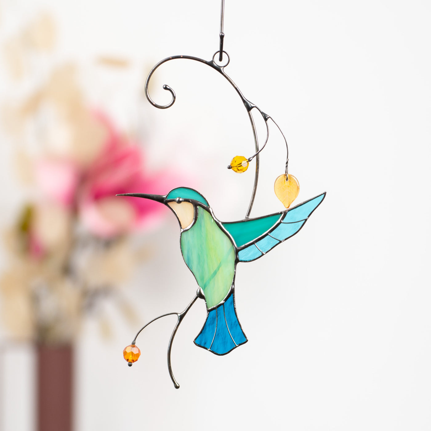 Delicate stained glass hummingbird with vibrant colors and beads, suspended in front of a blurred floral background.