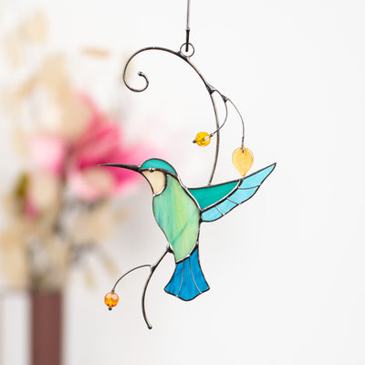 Delicate stained glass hummingbird with vibrant colors and beads, suspended in front of a blurred floral background.