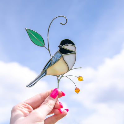 A hand holding a stained glass chickadee ornament under a bright blue sky with clouds.