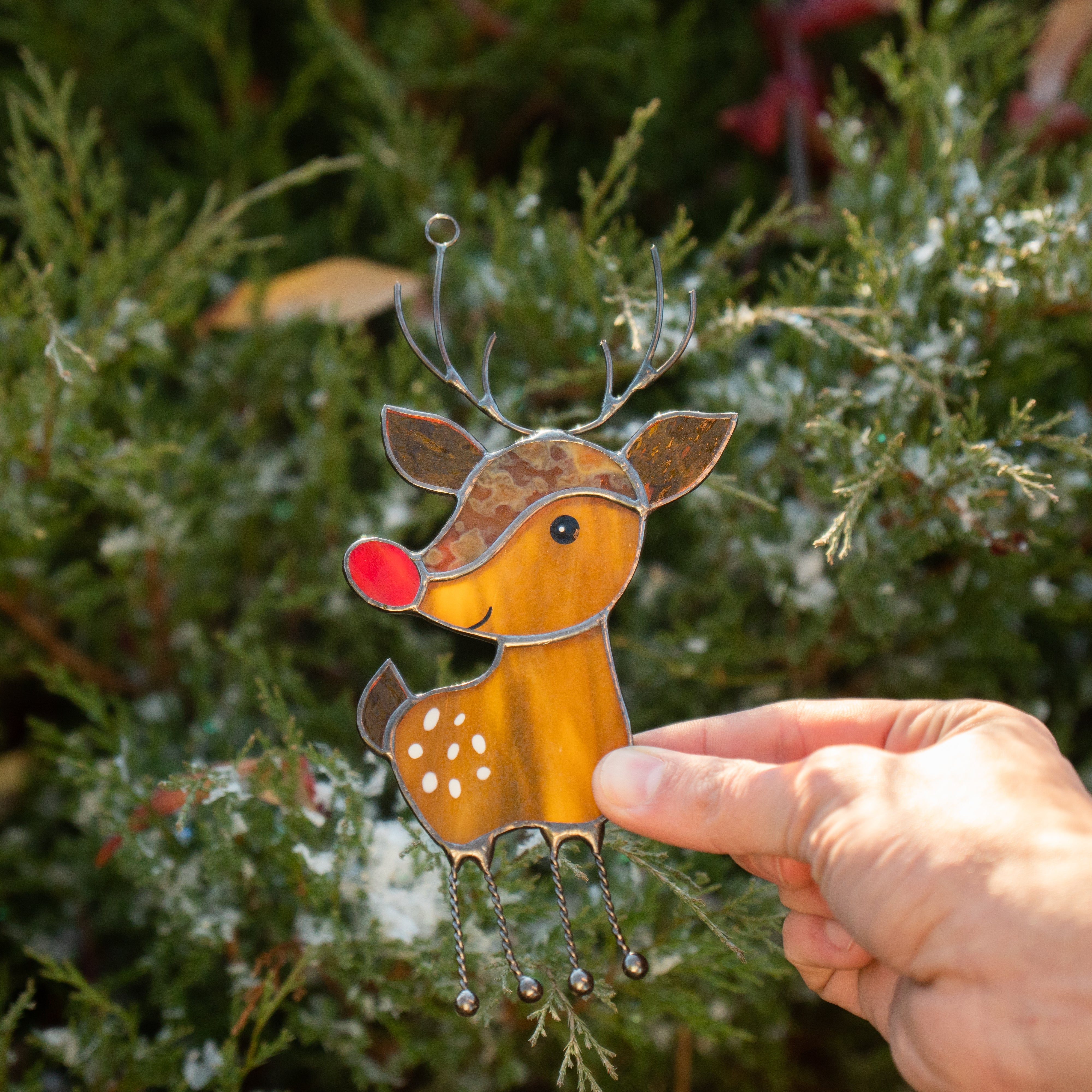 A hand holding a stained glass reindeer ornament with a red nose and twisted legs in front of evergreen branches.