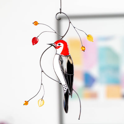 Stained glass woodpecker suncatcher hanging indoors with curved wire branches and red, orange, and yellow glass leaves.