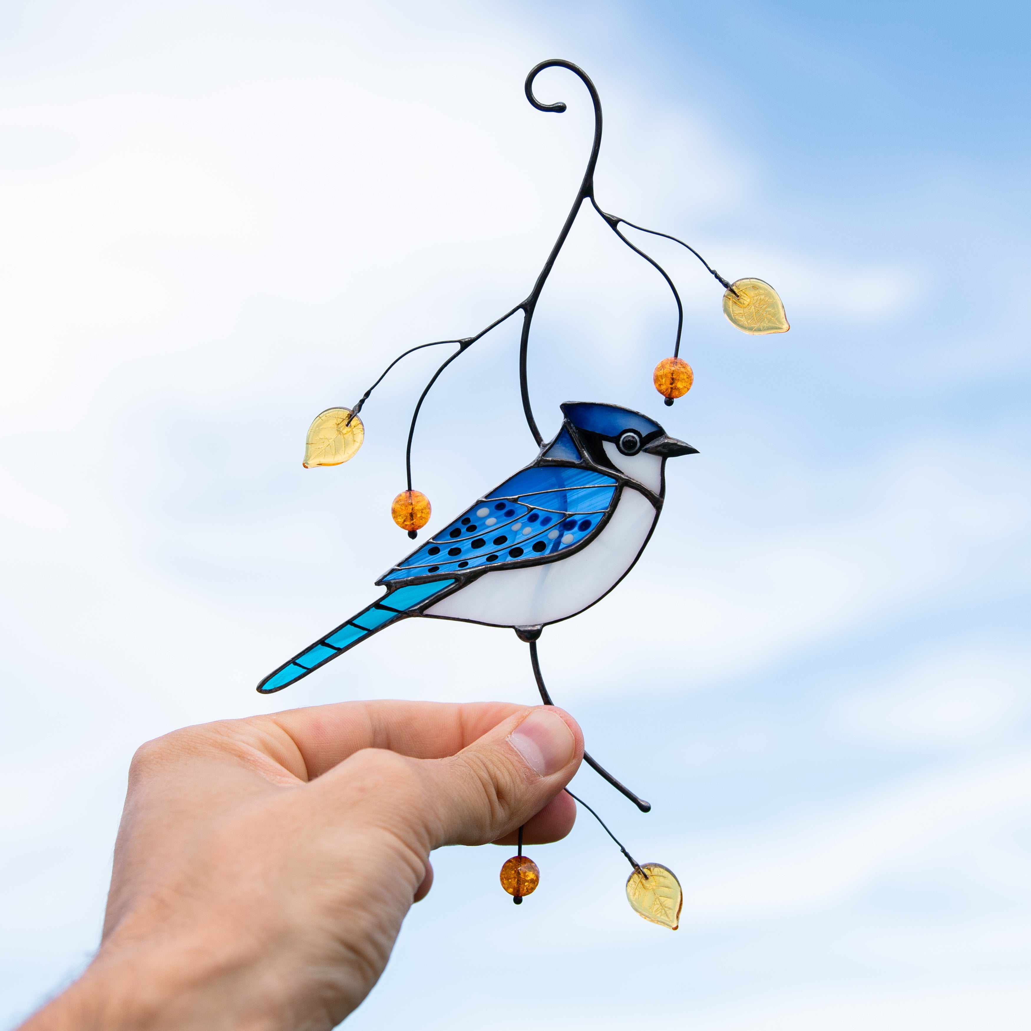 A hand holding a stained glass blue jay suncatcher with yellow leaves and beads against a bright sky