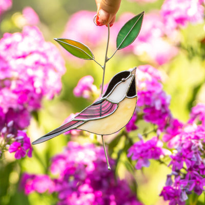 Hand holding stained glass chickadee with a semi-clear wing and green leaves, surrounded by blooming purple flowers