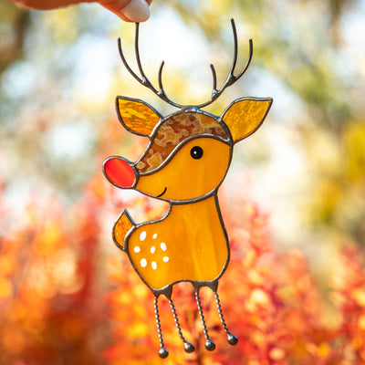 A stained glass reindeer ornament with a red nose and metal antlers is held up against warm orange autumn foliage.