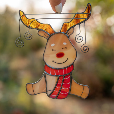 Smiling stained glass reindeer ornament with brown antlers and red scarf, held against a sunlit forest background.