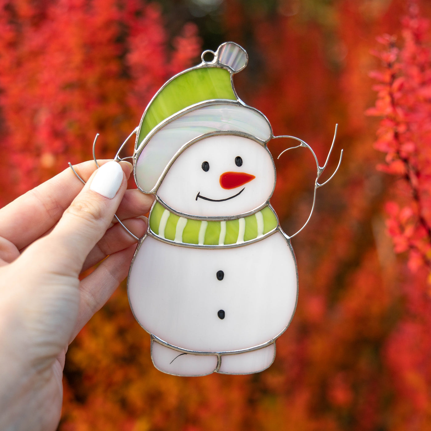 A stained glass snowman with a green hat and scarf, held by hand in front of red and orange background