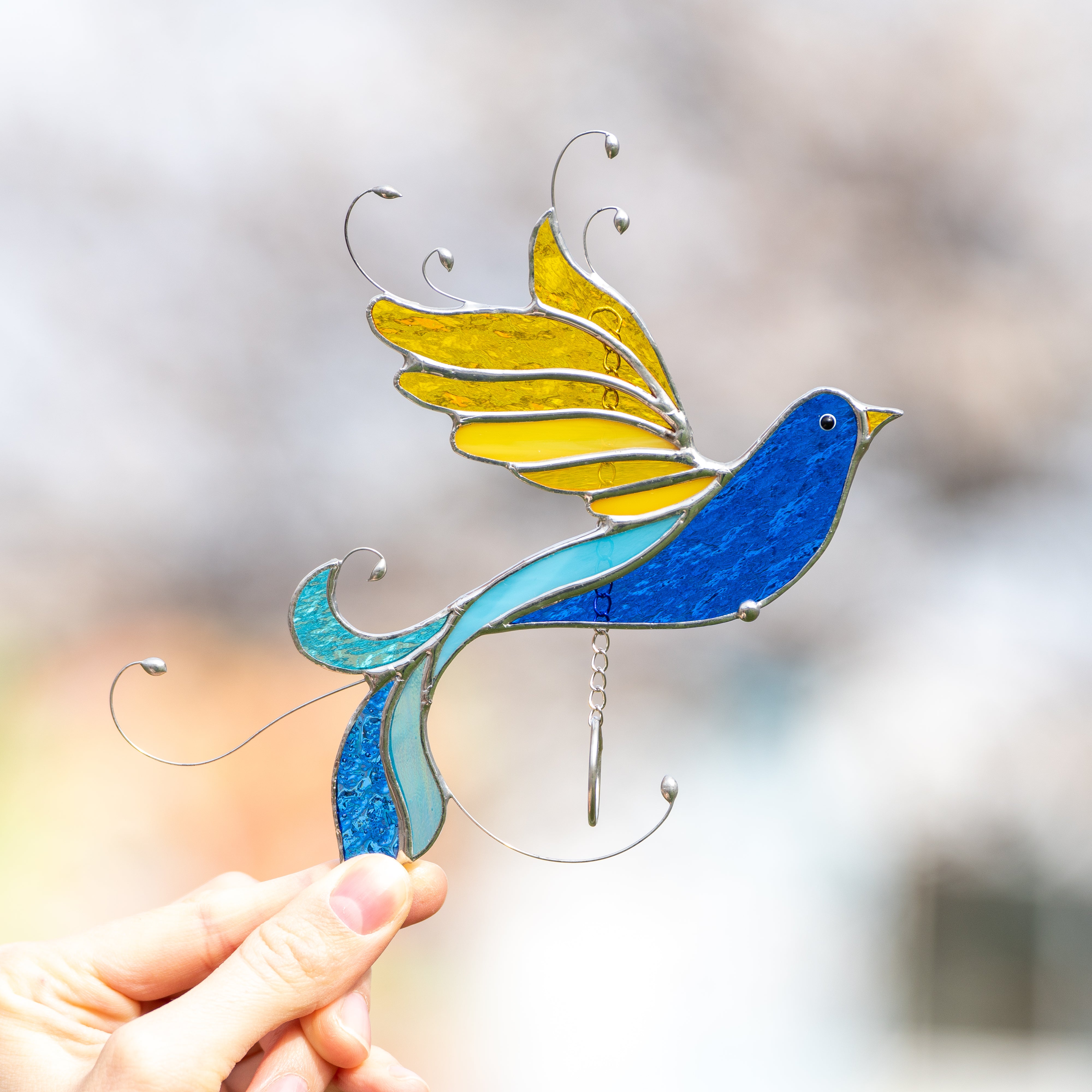 A stained glass bird in Ukrainian colors with yellow wings and curled blue tail, shown close-up in hand against a soft background.