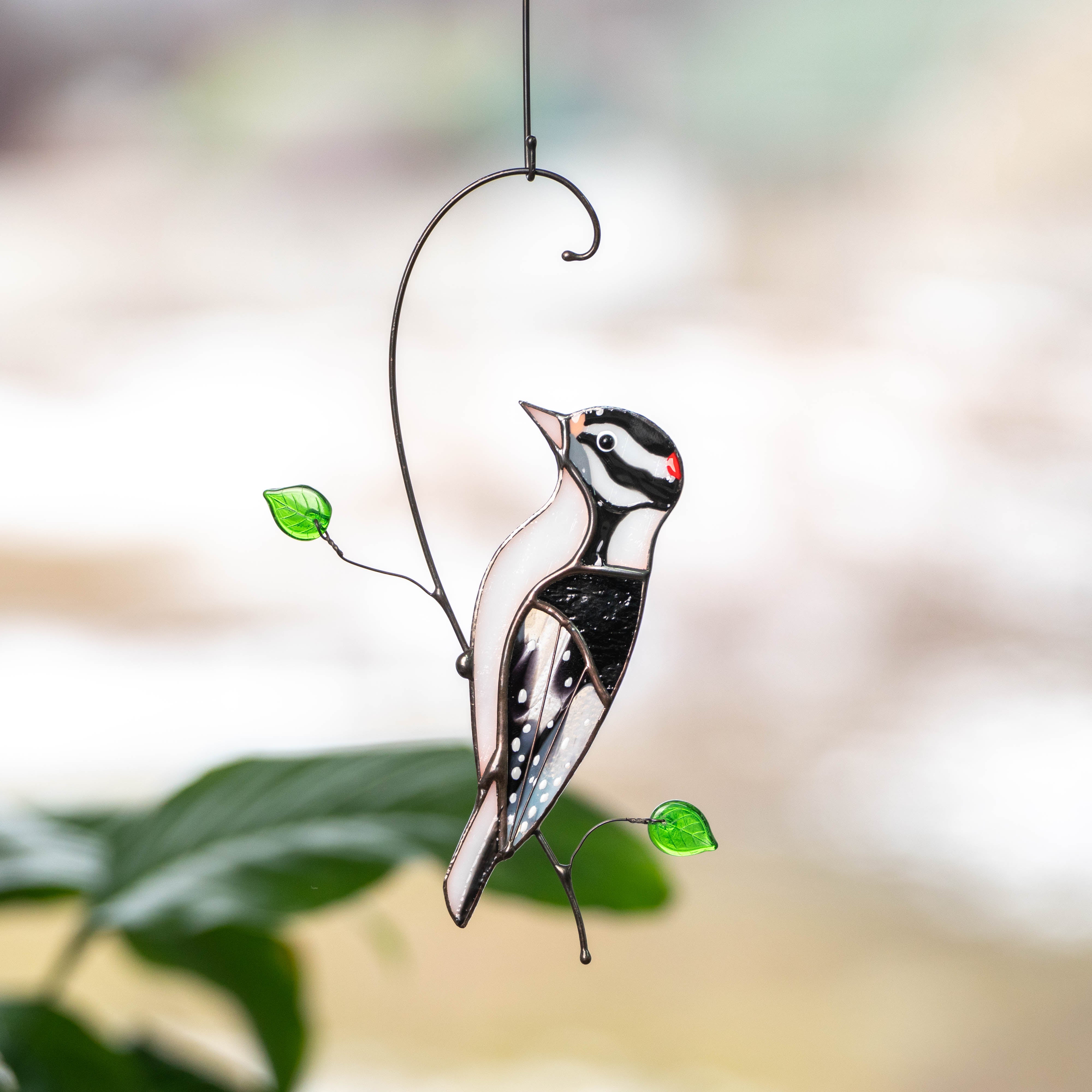 Black, white, and red stained glass downy woodpecker with green leaf accents hanging near a leafy background.