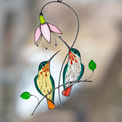 Stained glass green hummingbirds from orange and clear glass sitting on the branch with pink flower suncatcher