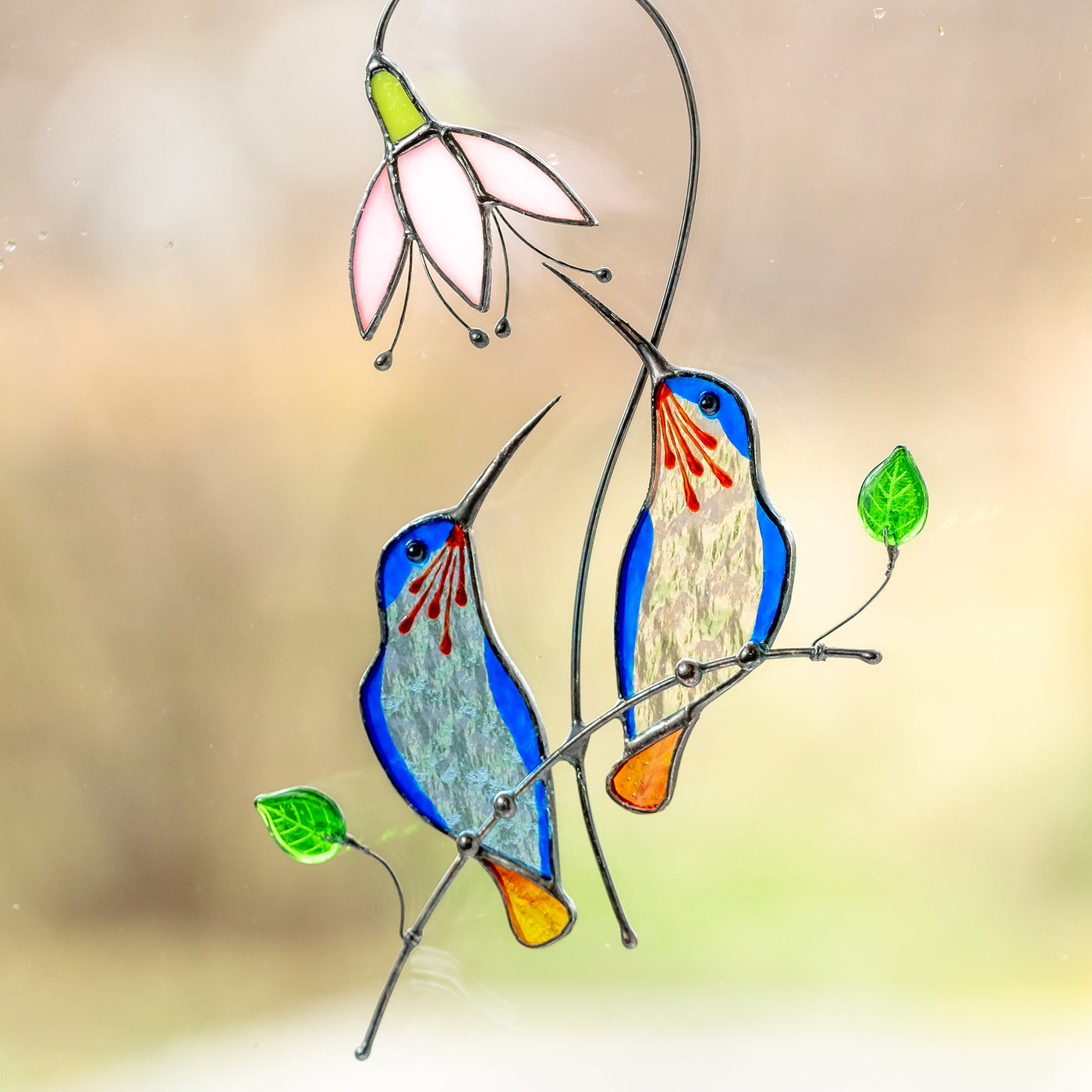Stained glass pair of hummingbirds from light blue and clear glass sitting on the branch with pink flower above suncatcher