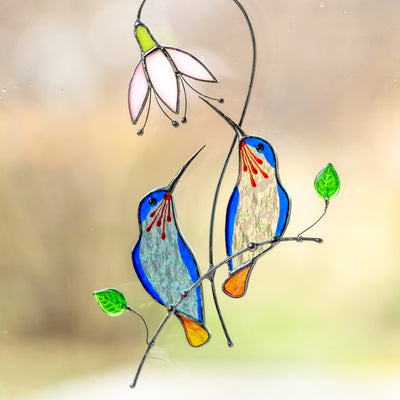 Stained glass pair of hummingbirds from light blue and clear glass sitting on the branch with pink flower above suncatcher